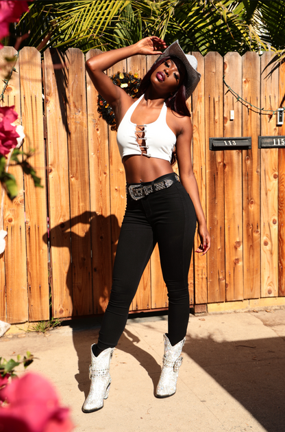 Woman in white top and black pants standing in front of a wooden fence with plants and flowers around.